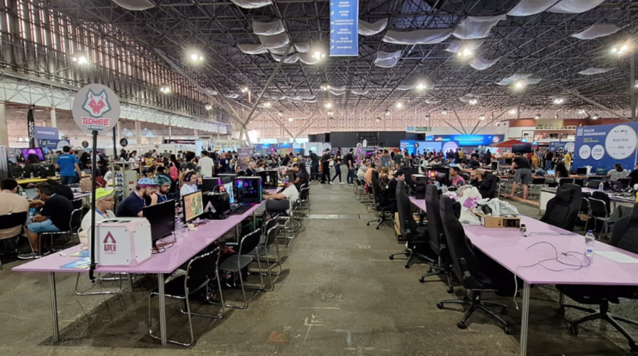 Evento de tecnologia Campus Party com muitos participantes em um espaço amplo, mesas com computadores e equipamentos de jogos.