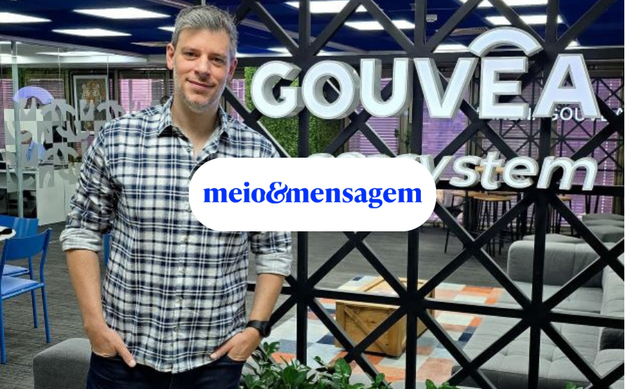 Michel Serebrinsky, novo CMDO da Gouvêa Ecosystem, está vestido de camisa xadrez e calça preta, na frente de uma estrutura de metal, com a logo da Gouvêa Ecosystem.